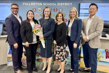 Dr. Gretchen Janson stands with five Fullerton School District trustees in a boardroom, holding a bouquet of flowers with a district sign and artwork in the background.