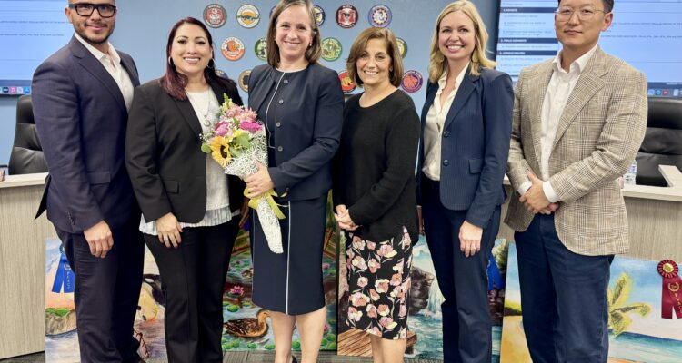 Dr. Gretchen Janson stands with five Fullerton School District trustees in a boardroom, holding a bouquet of flowers with a district sign and artwork in the background.