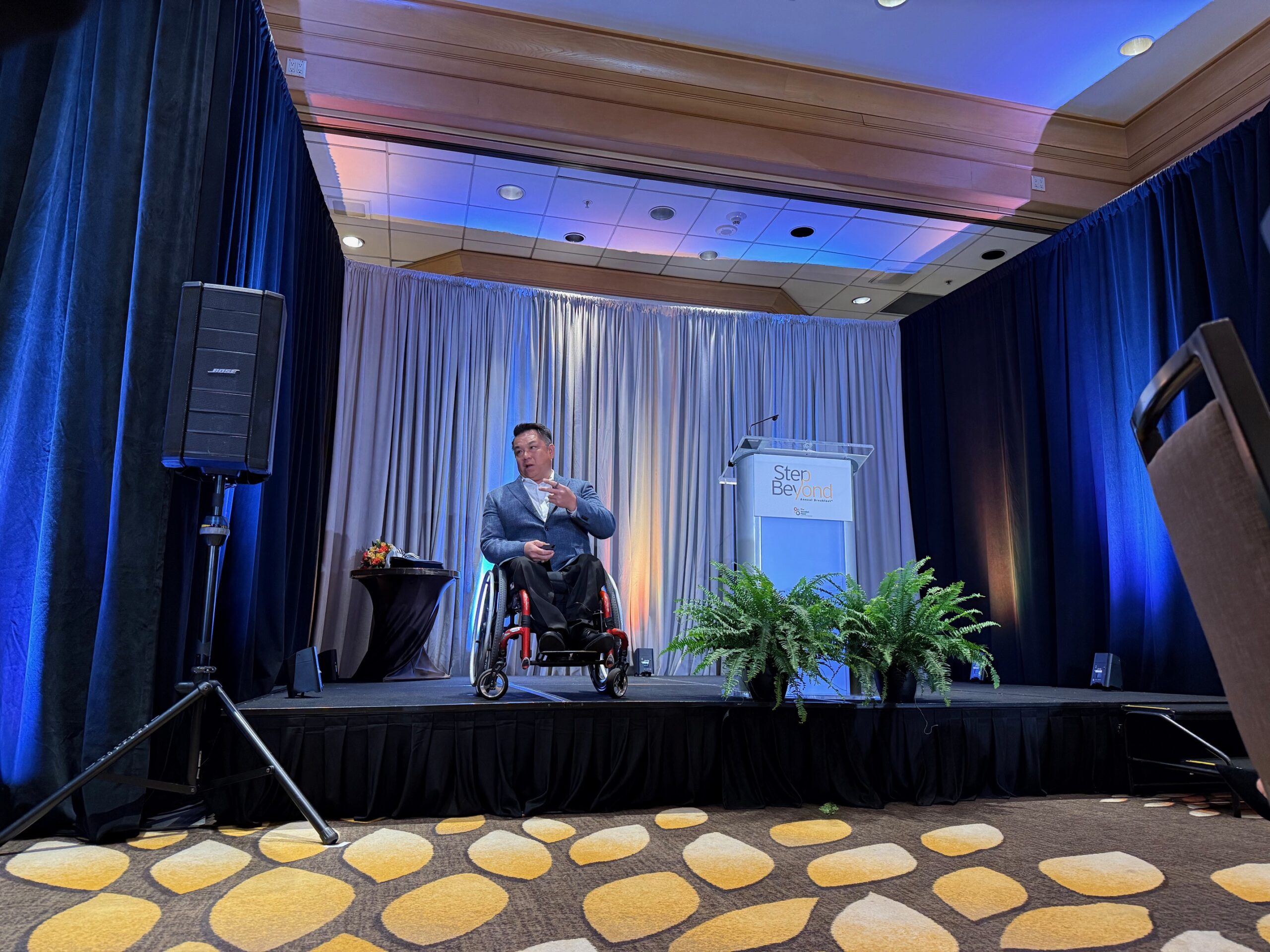 Dr. Stefan Bean speaks on a stage during The Wooden Floor’s Step Beyond Annual Breakfast, with blue lighting, curtains and a podium in the background.