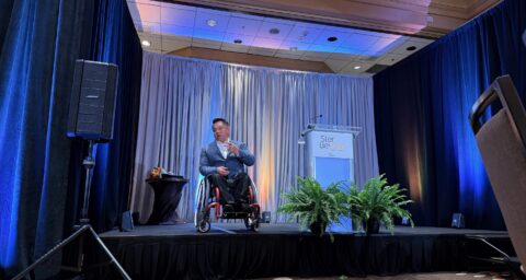 Dr. Stefan Bean speaks on a stage during The Wooden Floor’s Step Beyond Annual Breakfast, with blue lighting, curtains and a podium in the background.