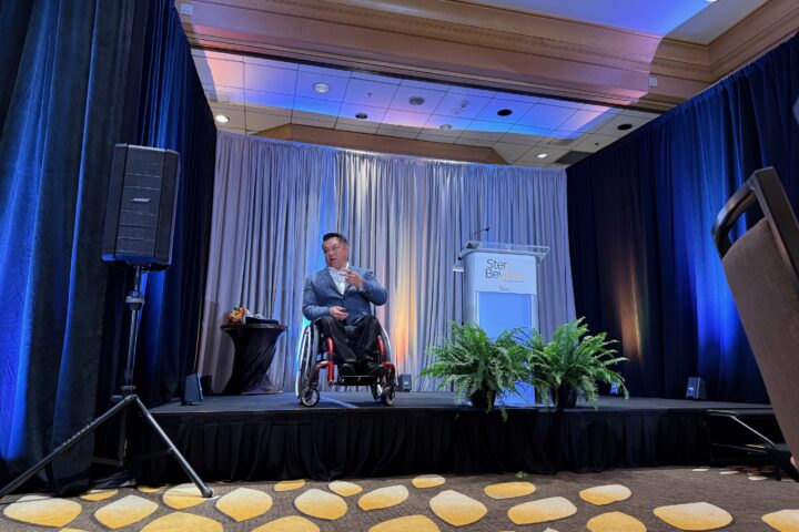 Dr. Stefan Bean speaks on a stage during The Wooden Floor’s Step Beyond Annual Breakfast, with blue lighting, curtains and a podium in the background.