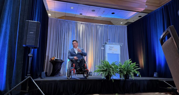 Dr. Stefan Bean speaks on a stage during The Wooden Floor’s Step Beyond Annual Breakfast, with blue lighting, curtains and a podium in the background.