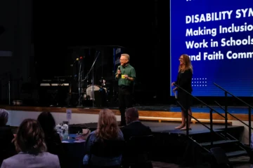 Dr. Jeff Hittenberger speaks on stage at the Disability Symposium while attendees listen from round tables.