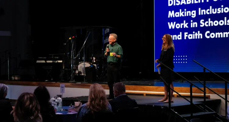 Dr. Jeff Hittenberger speaks on stage at the Disability Symposium while attendees listen from round tables.