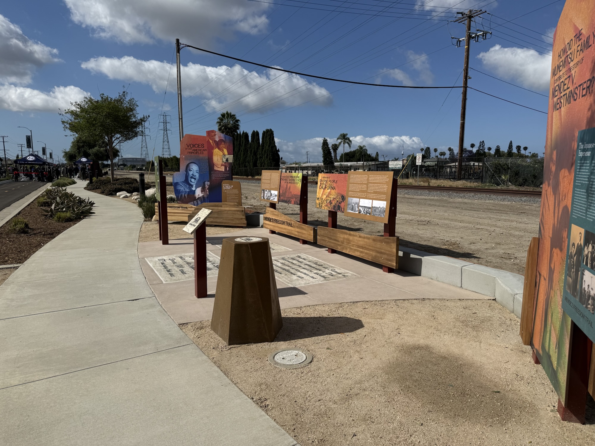 Interpretive panels and seating along the Mendez Freedom Trail in Westminster, with the dedication ceremony visible in the distance along Hoover Street.