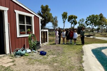 A group of elementary students and their principal stand near a red barn and garden area on Golden View Elementary’s school farm.