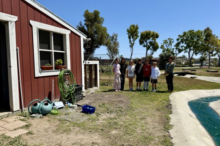 A group of elementary students and their principal stand near a red barn and garden area on Golden View Elementary’s school farm.