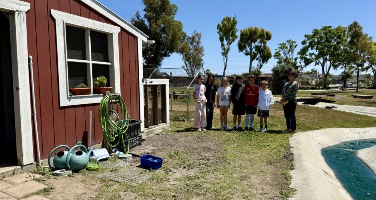 A group of elementary students and their principal stand near a red barn and garden area on Golden View Elementary’s school farm.
