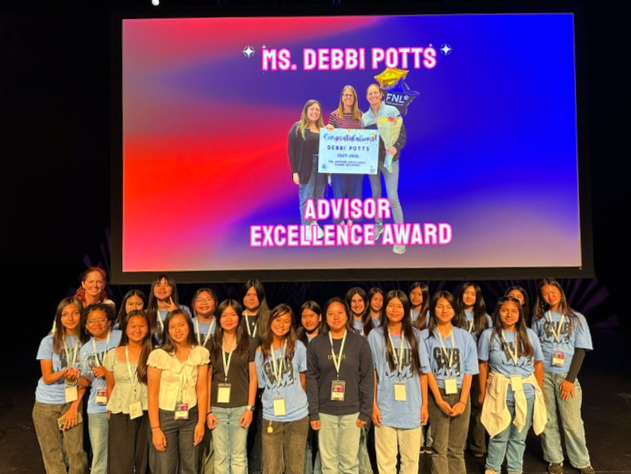 Debbi Potts stands with a group of students onstage beneath a screen recognizing her as the recipient of the Advisor Excellence Award at the Friday Night Live ceremony.