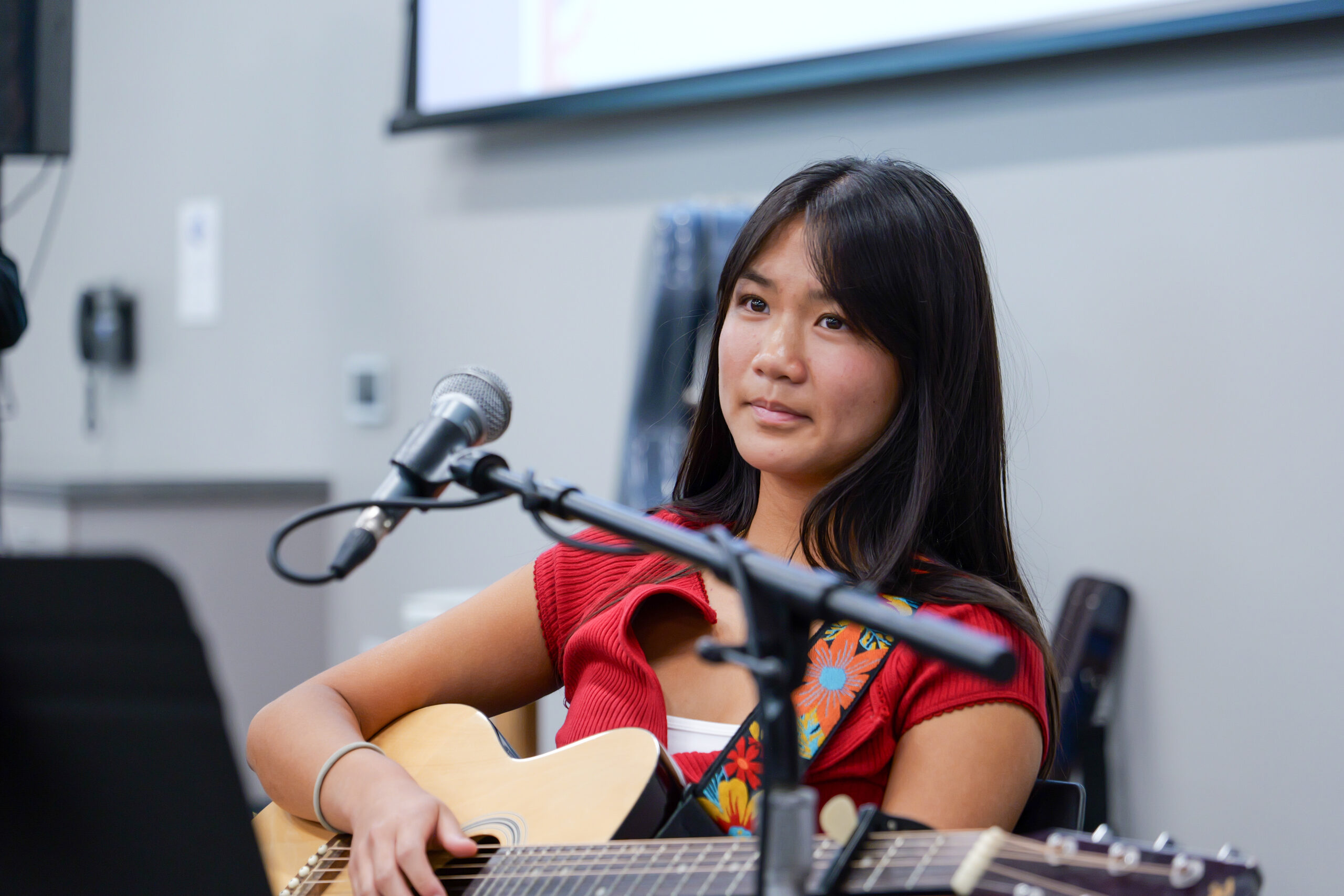 Ella Wu holds an acoustic guitar while seated near a microphone during the USA 250-OC Theme Song Competition showcase.