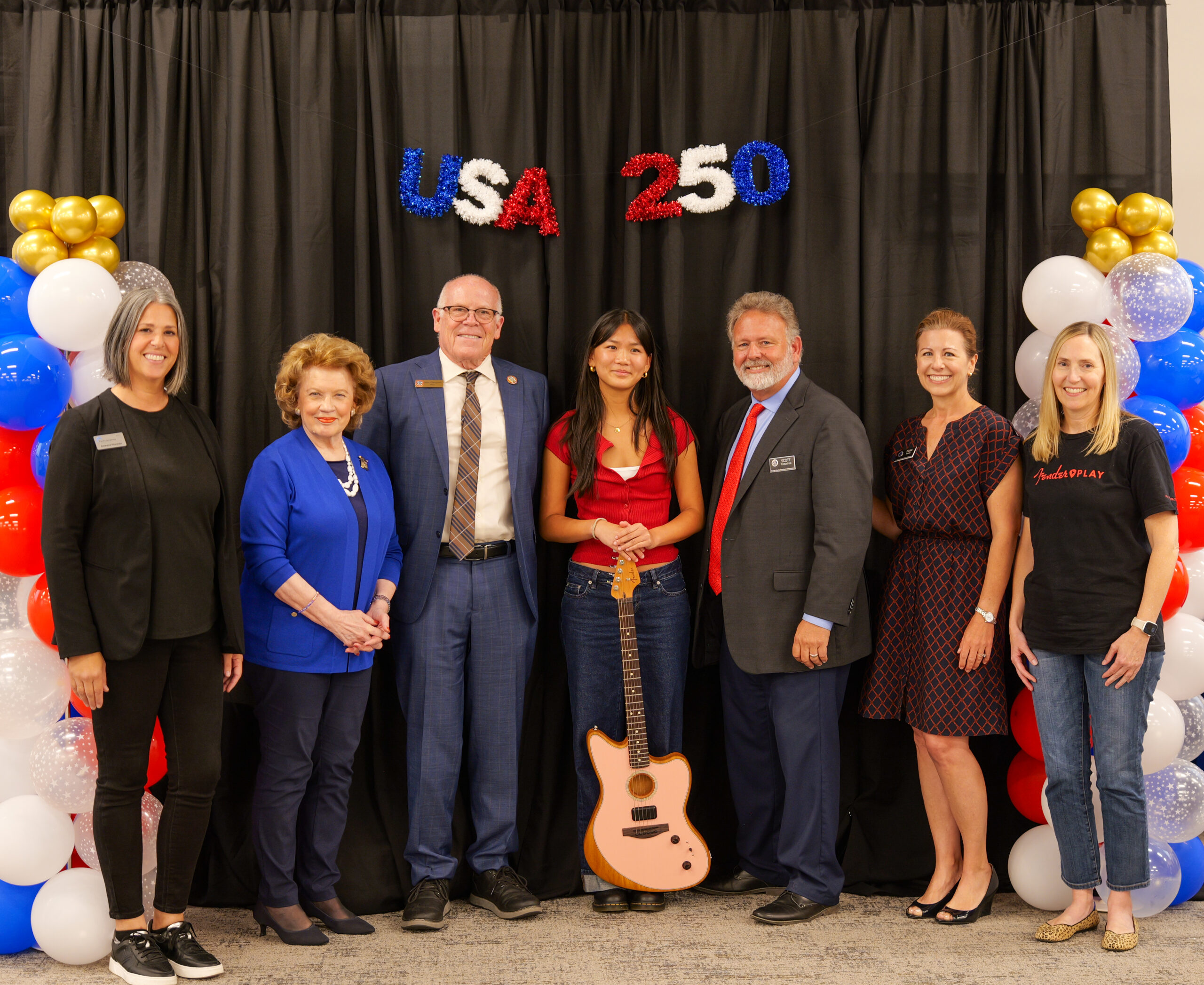 USA 250-OC Theme Song Competition grand prize winner Ella Wu stands with community leaders and program partners while holding a guitar in front of a USA 250 backdrop following the June 6 awards ceremony.