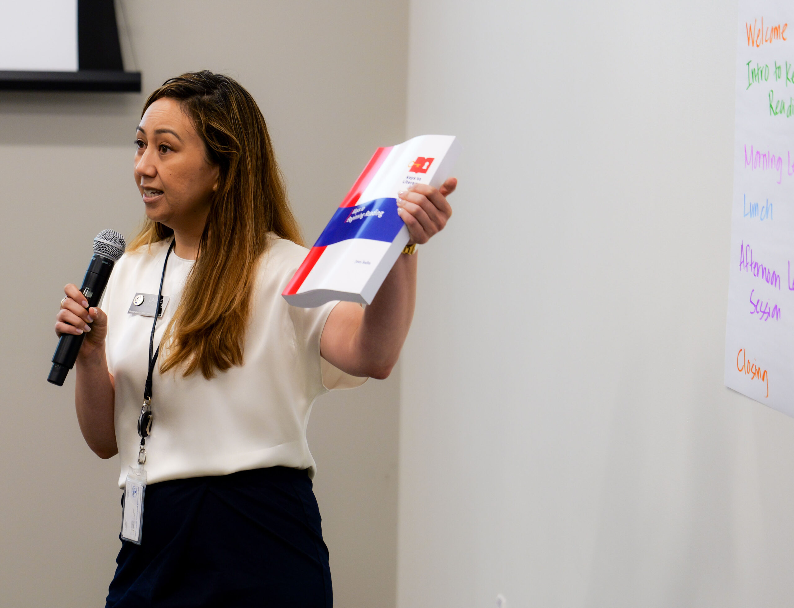 Dr. Laurie Virtusio holds up a training book while speaking into a microphone during a literacy training session at the OCDE Conference Center.