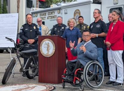 Orange County Superintendent of Schools Dr. Stefan Bean speaks from his wheelchair at an e-bike safety event, with law enforcement, health and community partners gathered behind him.