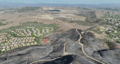 Aerial view of charred, blackened hillsides and winding dirt roads following the Norte Fire in San Clemente, with residential neighborhoods bordering the burned area and rolling hills in the distance.