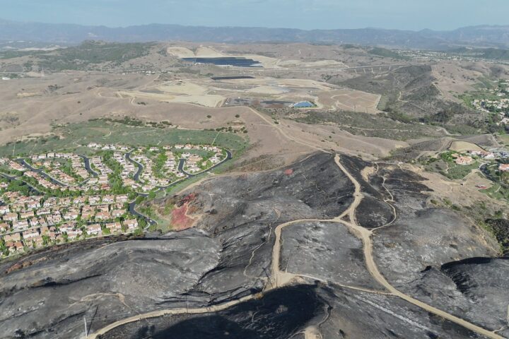 Aerial view of charred, blackened hillsides and winding dirt roads following the Norte Fire in San Clemente, with residential neighborhoods bordering the burned area and rolling hills in the distance.