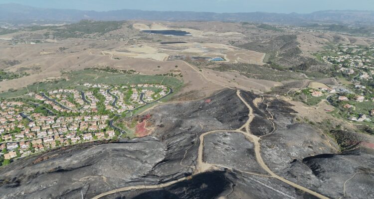 Aerial view of charred, blackened hillsides and winding dirt roads following the Norte Fire in San Clemente, with residential neighborhoods bordering the burned area and rolling hills in the distance.