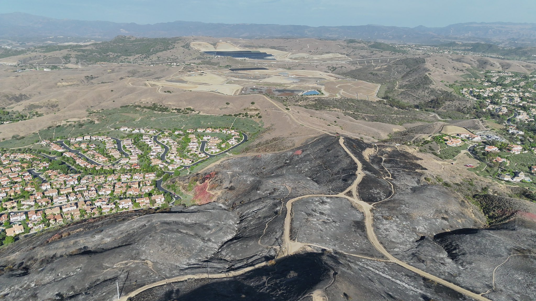 Aerial view of charred, blackened hillsides and winding dirt roads following the Norte Fire in San Clemente, with residential neighborhoods bordering the burned area and rolling hills in the distance.
