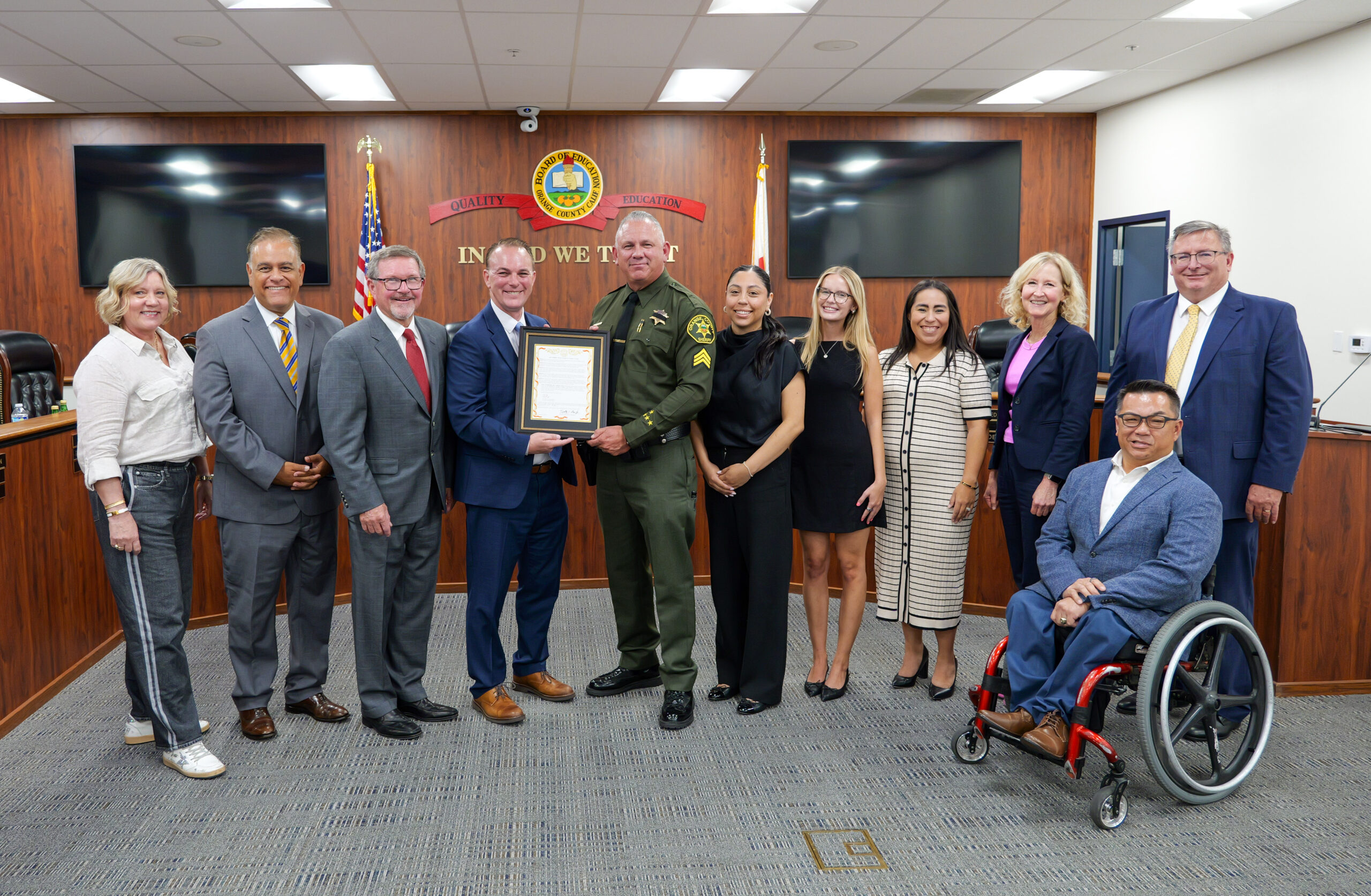 Ten people pose for a group photo in the Orange County Board of Education meeting room, with a uniformed sheriff's sergeant and a man in a blue suit holding a framed Red Ribbon Week resolution at the center.