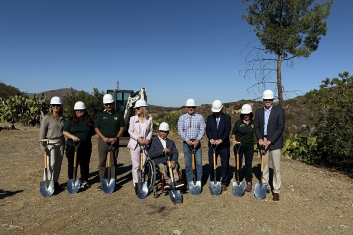 A group of OCDE and Orange County Board of Education leaders, Inside the Outdoors staff and project partners wear white hard hats and pose with shovels at the Rancho Soñado groundbreaking site, with hills and construction equipment in the background.