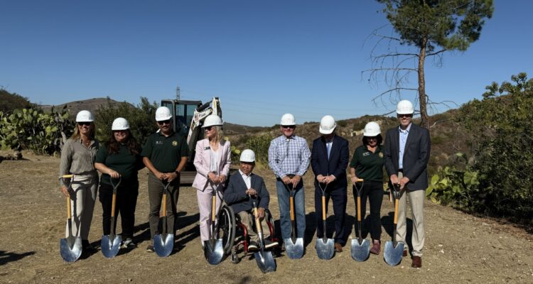 A group of OCDE and Orange County Board of Education leaders, Inside the Outdoors staff and project partners wear white hard hats and pose with shovels at the Rancho Soñado groundbreaking site, with hills and construction equipment in the background.