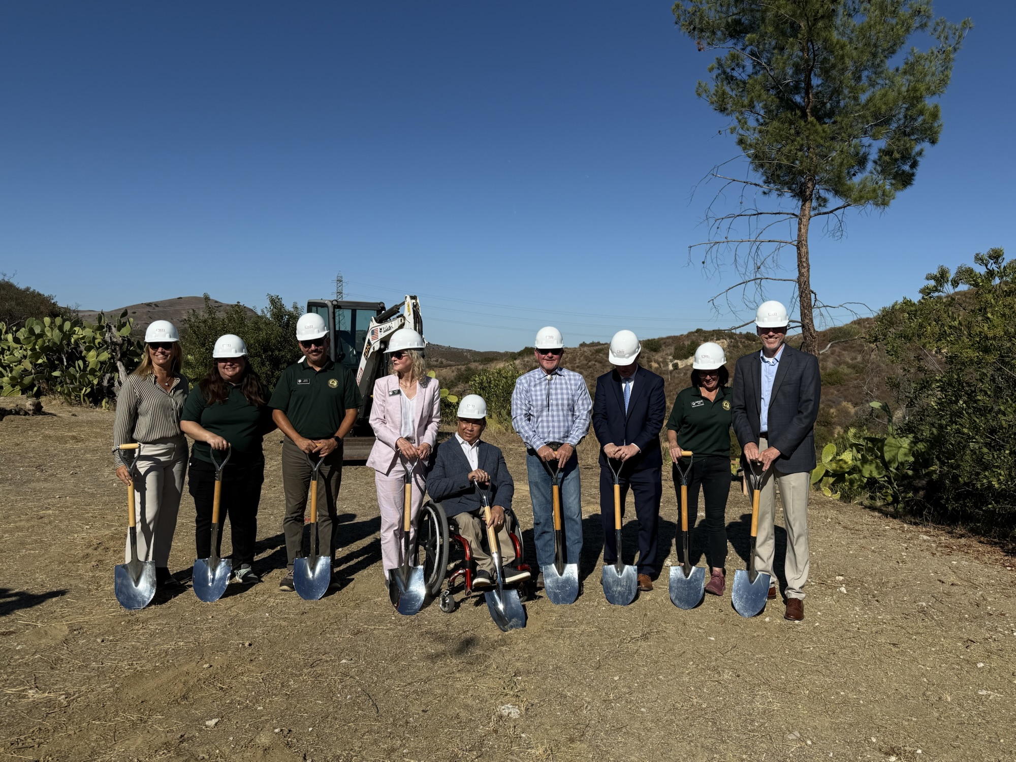A group of OCDE and Orange County Board of Education leaders, Inside the Outdoors staff and project partners wear white hard hats and pose with shovels at the Rancho Soñado groundbreaking site, with hills and construction equipment in the background.